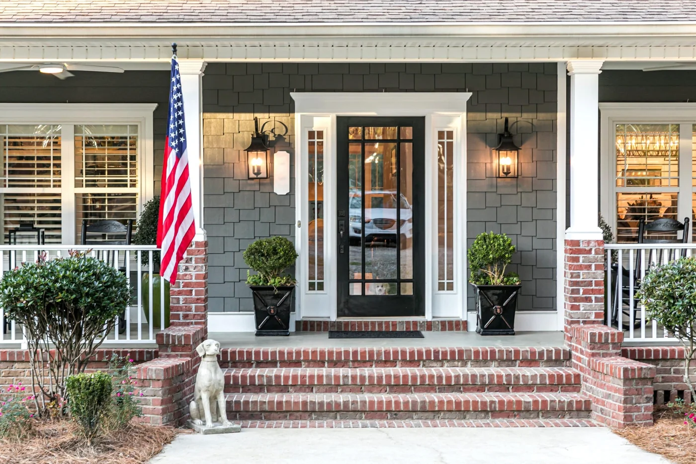 Front Door Entrance To A Large Two Story Blue Gray House With Wood And Vinyl Siding And A Large American Flag.
