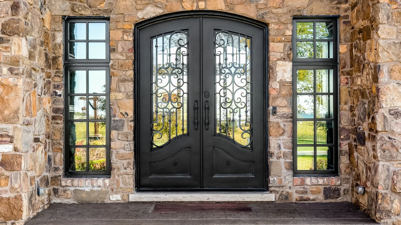 There is a dark brown door with a beautiful iron design. There is also glass in the center of the door and windows on either side.
