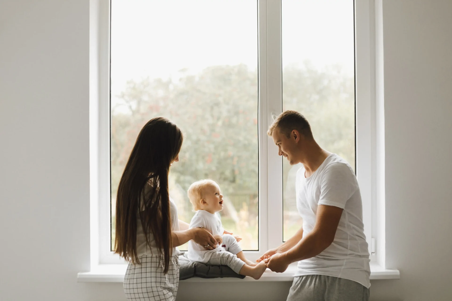 A young family plays in front of a newly replaced window. The father sets the baby on the windowsill to enjoy the view.