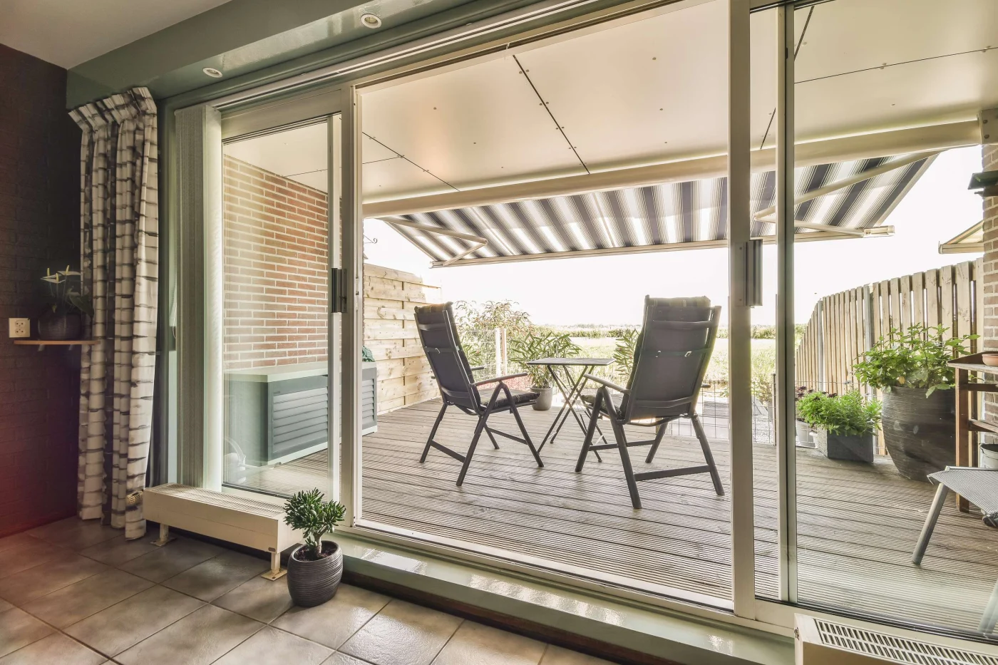 A stunning, wide sliding patio door with retracted blinds looking out on a brick-enclosed patio with a small table and two chairs, and a striped overhead canopy.
