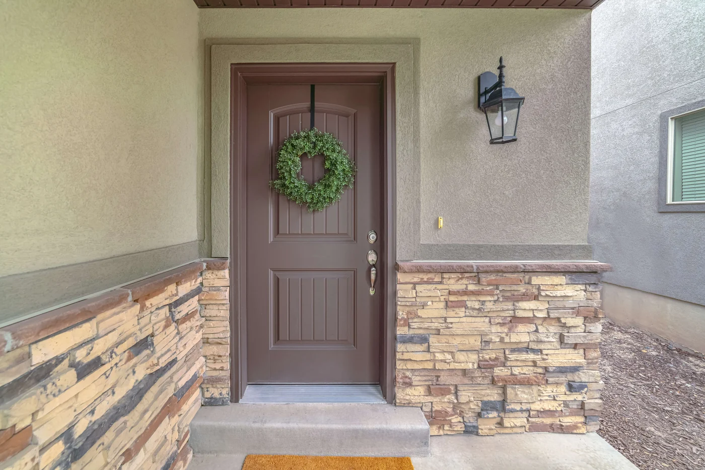 A dark brown front door with a wreath on a green stucco house with a stone base